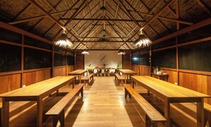 A restaurant with wooden tables and benches, a thatched roof, and a wall with a circular emblem. - Amazon Field Station Byinkaterra (Tambopata)