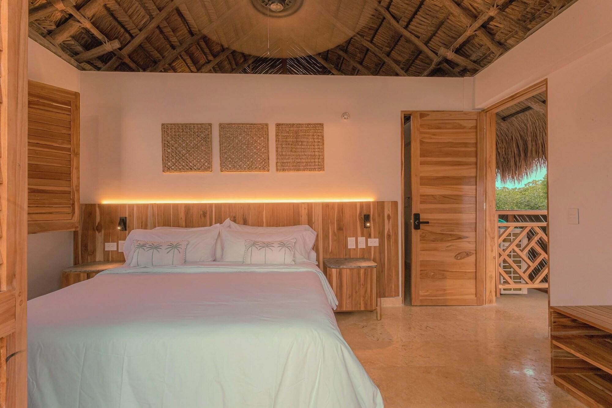 A bedroom with a wooden headboard, a bed with white bedding, two wall-mounted woven panels, and a wooden door leading to a balcony.