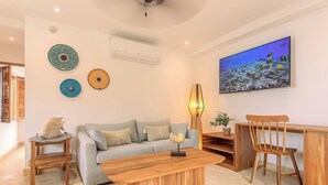 Un salon avec un canapé gris, une table basse en bois et deux chaises en bois. Des assiettes décoratives ornent le mur, et un grand écran de télévision diffuse une scène sous-marine.