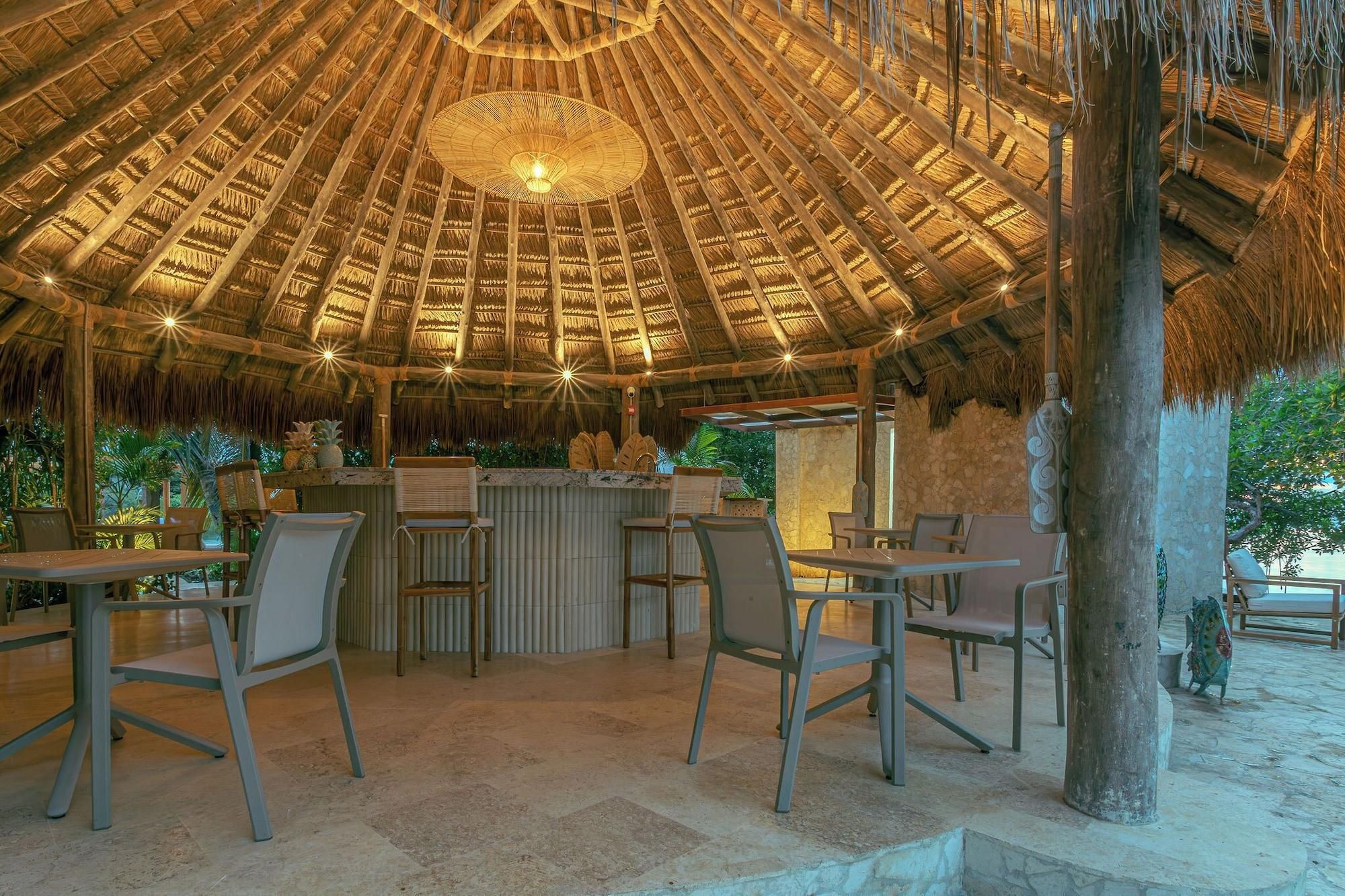An outdoor dining area with a thatched roof, stone flooring, and several tables and chairs.