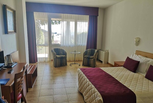 A hotel room with a bed, a desk, two chairs, a small table, and a window with curtains. - Relax Hotel Acquaviva - Tuscany Hills (Casole d'Elsa)