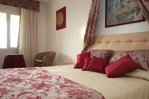 A hotel room with a large bed, red and beige bedding, a chair, a table, and a painting on the wall. - Relax Hotel Acquaviva - Tuscany Hills (Casole d'Elsa)