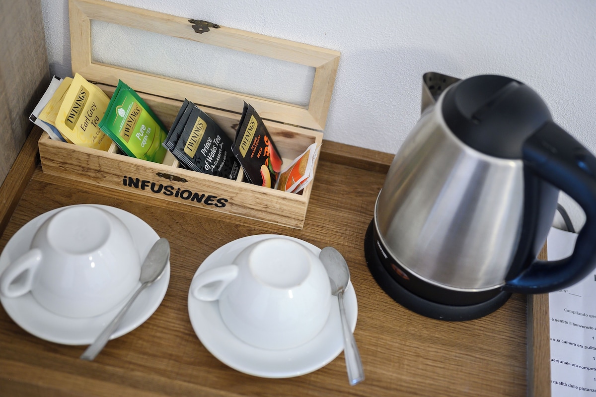 A wooden tray with a stainless steel kettle, two white cups with saucers, and a box of Twinings tea bags.