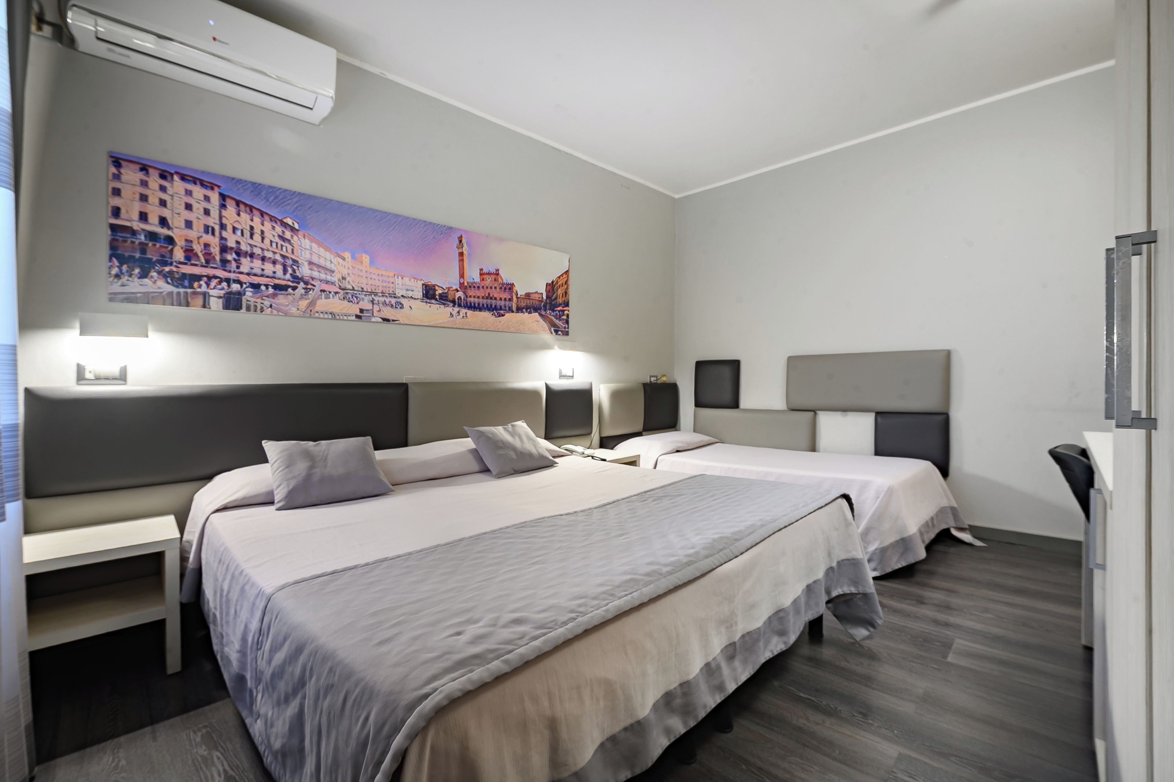 Camera d'albergo con due letti, un quadro di un panorama cittadino e una scrivania con una sedia.