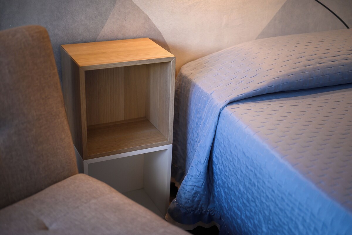 A bedroom with a wooden bedside table, a blue quilted bedspread, and a grey upholstered chair.