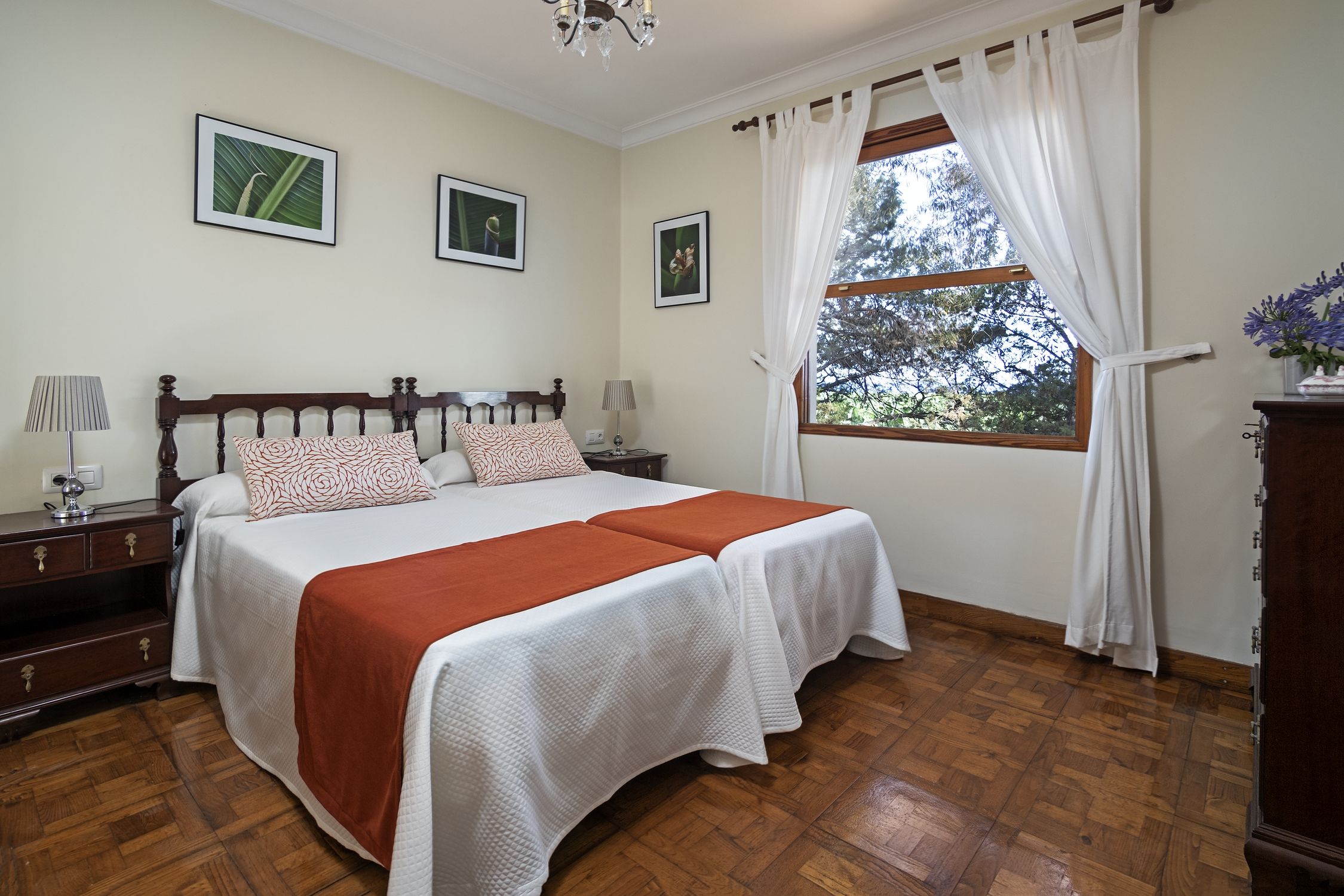 A bedroom with a wooden bed, two bedside tables, a nightstand, a window with curtains, and framed pictures on the wall.