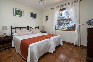 Un dormitorio con una cama de madera, dos mesitas de noche, una mesita auxiliar, una ventana con cortinas y cuadros enmarcados en la pared.