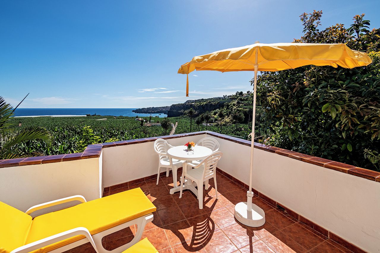 En balkon med et bord, stole og en gul parasol, samt udsigt over havet.