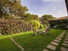 Un jardín con una mesa y sillas, un seto y un edificio con tejado de tejas.