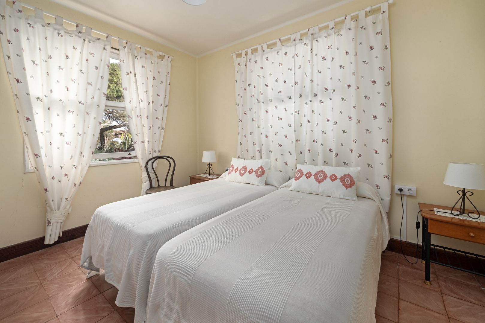 A room with two beds, a chair, a table with a lamp, and a window with curtains.