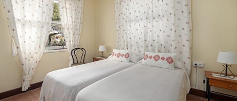 A room with two beds, a chair, a table with a lamp, and a window with curtains.