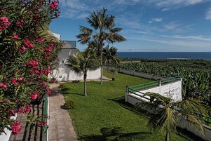 Un jardín bien cuidado con palmeras y plantas con flores, un edificio blanco con balcón, y vista al océano.