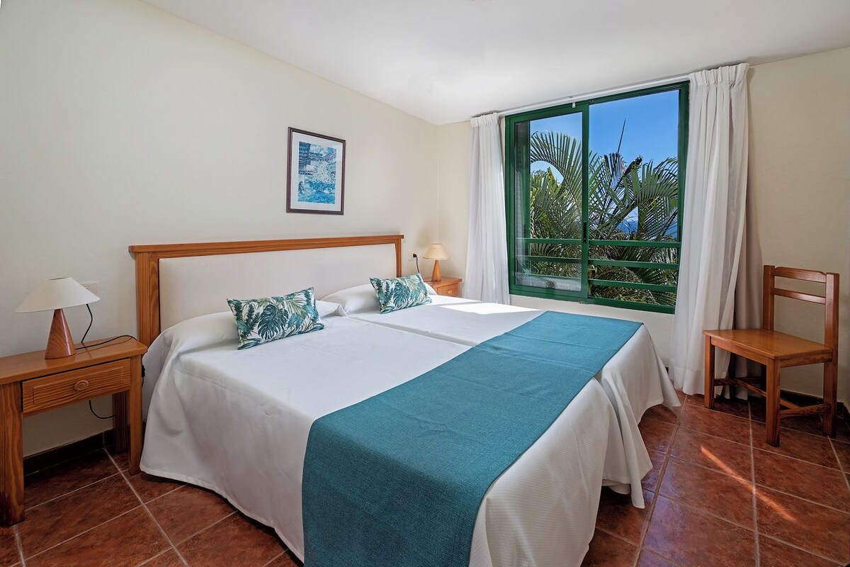 A hotel room with a large bed, a desk, a chair, a window with a view of palm trees, and a framed picture on the wall.