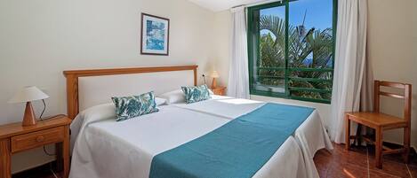 A hotel room with a large bed, a desk, a chair, a window with a view of palm trees, and a framed picture on the wall.