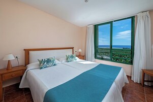 Una cama doble con una manta azul, dos almohadas, una mesita de noche con una lámpara y una ventana con vistas al mar.