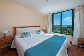 Un letto matrimoniale con una coperta blu, due cuscini, un comodino con una lampada e una finestra vista mare.