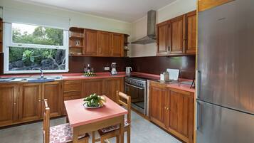 A kitchen with wooden cabinets, a stainless steel refrigerator, a window with a view of greenery, and a table with a bowl of fruit.