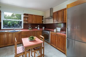 A kitchen with wooden cabinets, a stainless steel refrigerator, a window with a view of greenery, and a table with a bowl of fruit. - Sabinas & Terrazas Malpaís (Icod de los Vinos)