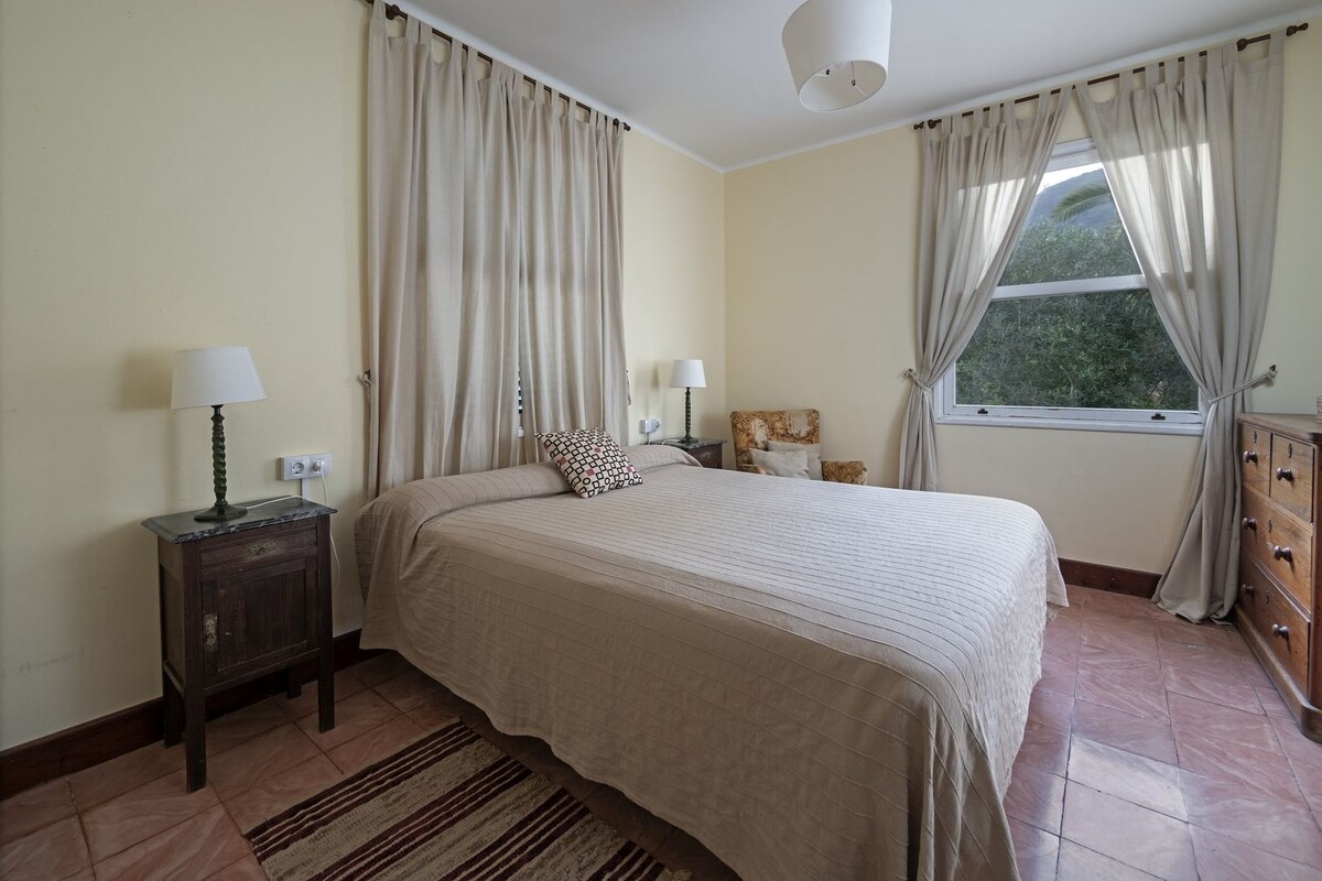 A bedroom with a bed, two bedside tables with lamps, a dresser, a window with curtains, and a view of greenery outside.