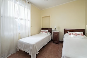 Habitación de hotel con dos camas individuales, una mesita de noche con lámpara y una ventana con cortinas traslúcidas.