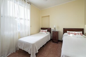 Habitación de hotel con dos camas individuales, una mesita de noche con lámpara y una ventana con cortinas traslúcidas.