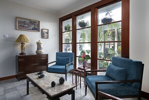 Una habitación con un ventanal grande, dos sillones azules, una mesita con adornos, un aparador y un jarrón con flores.