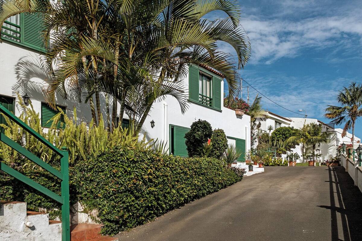 A white building with green shutters, a paved road, and a well-maintained hedge.