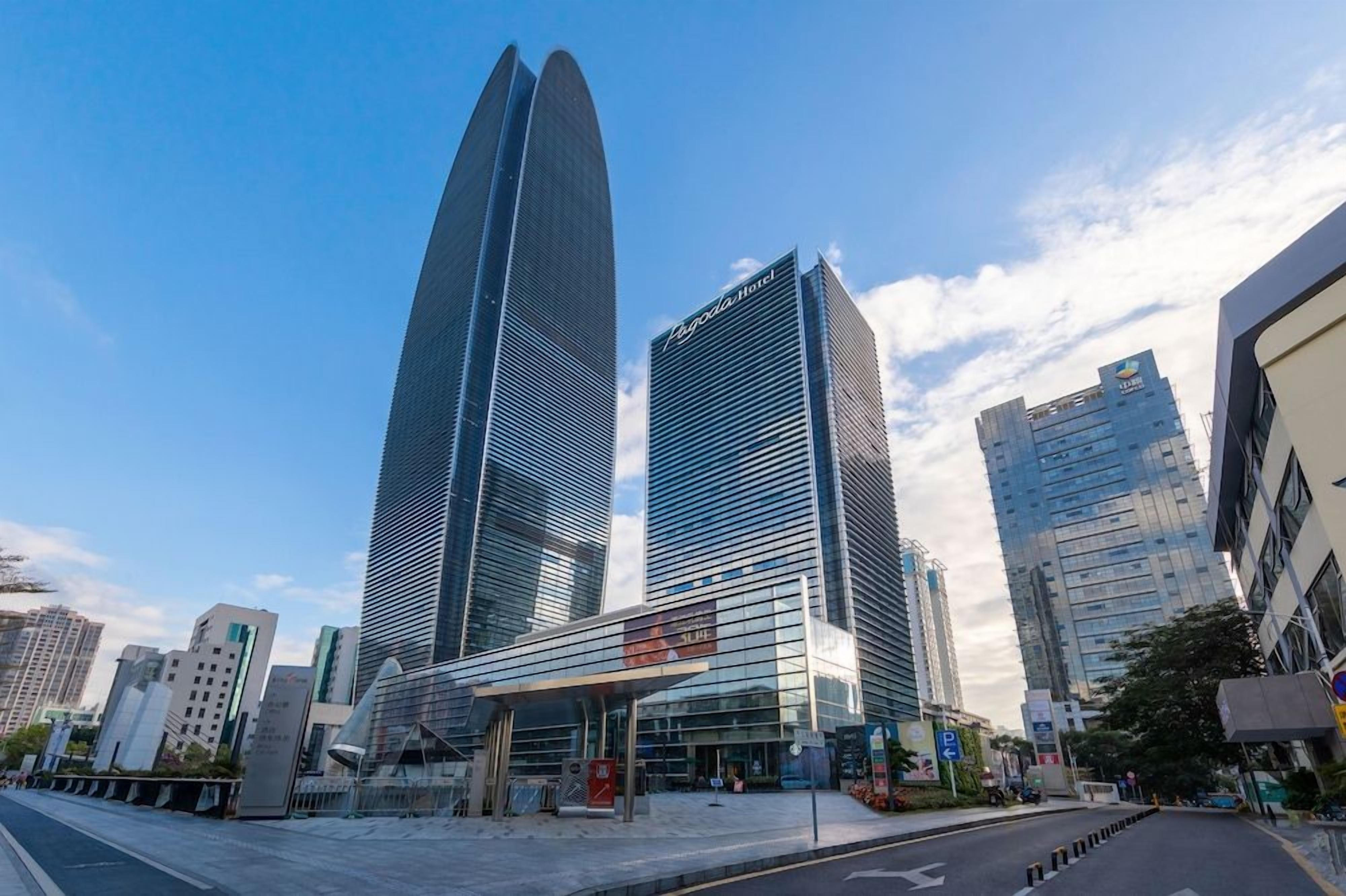 A modern hotel with a prominent glass facade, situated in a cityscape with high-rise buildings.