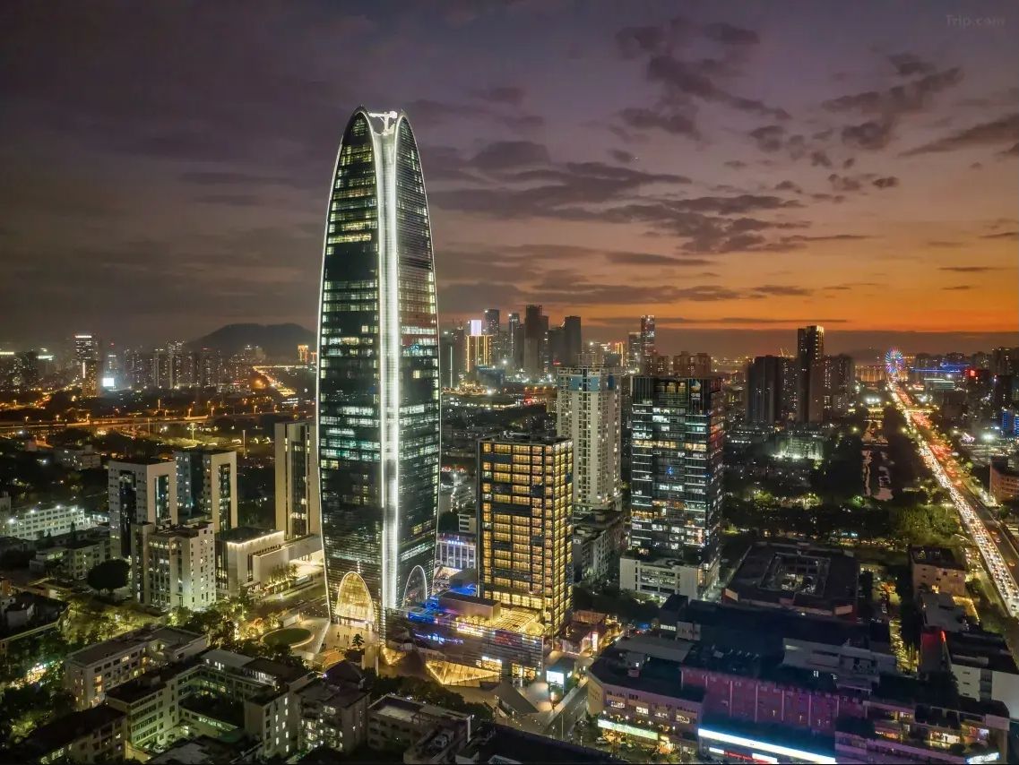 Paisagem urbana noturna com um arranha-céu curvo imponente, rodeado por outros edifícios altos e ruas iluminadas.