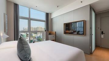 A modern hotel room with a large bed, a desk, a mirror, and a view of the cityscape.