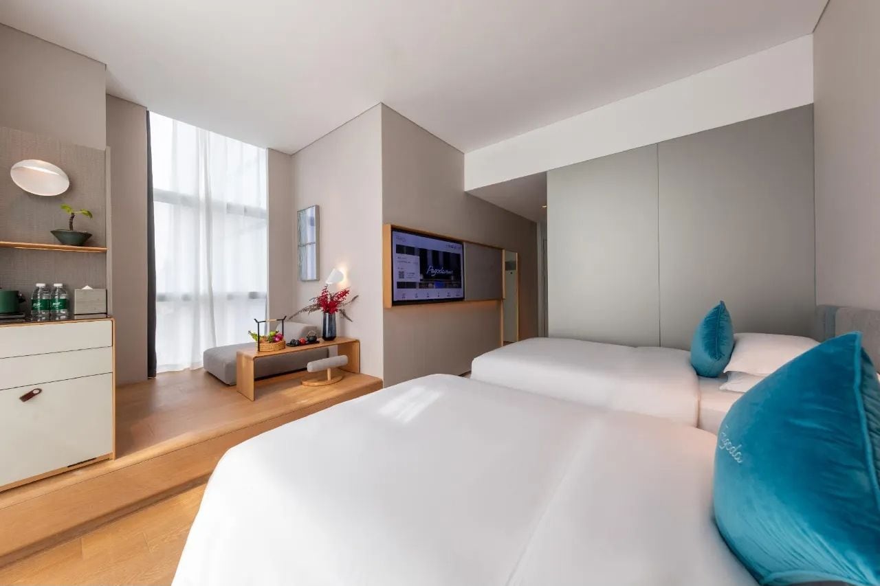 A modern hotel room with two beds, a desk with a computer, a TV, and a window with sheer curtains.
