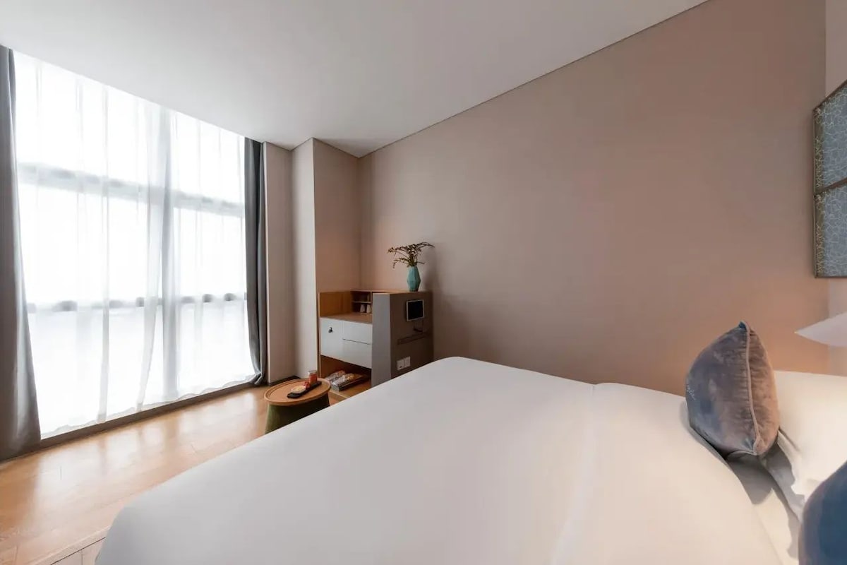 A hotel room with a large bed, a bedside table, a small desk, and a window with curtains.