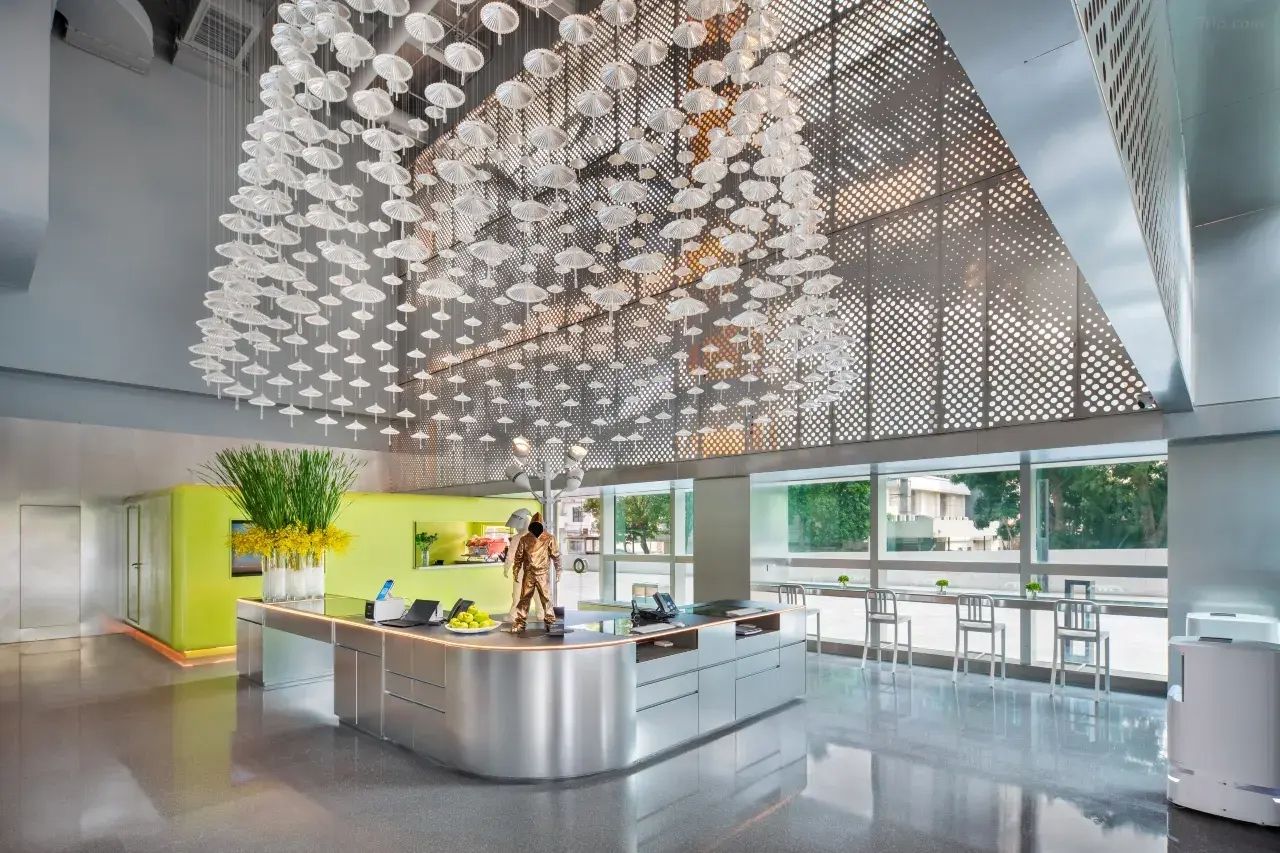A modern hotel lobby with a central reception desk, large windows, and a ceiling adorned with hanging white umbrellas.