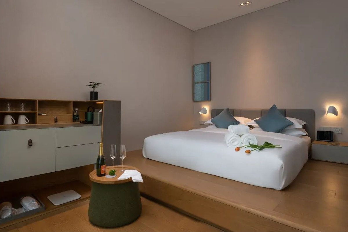 A modern hotel room with a large bed, bedside tables, a vanity with a mirror, and a small round table with a bottle and glasses.