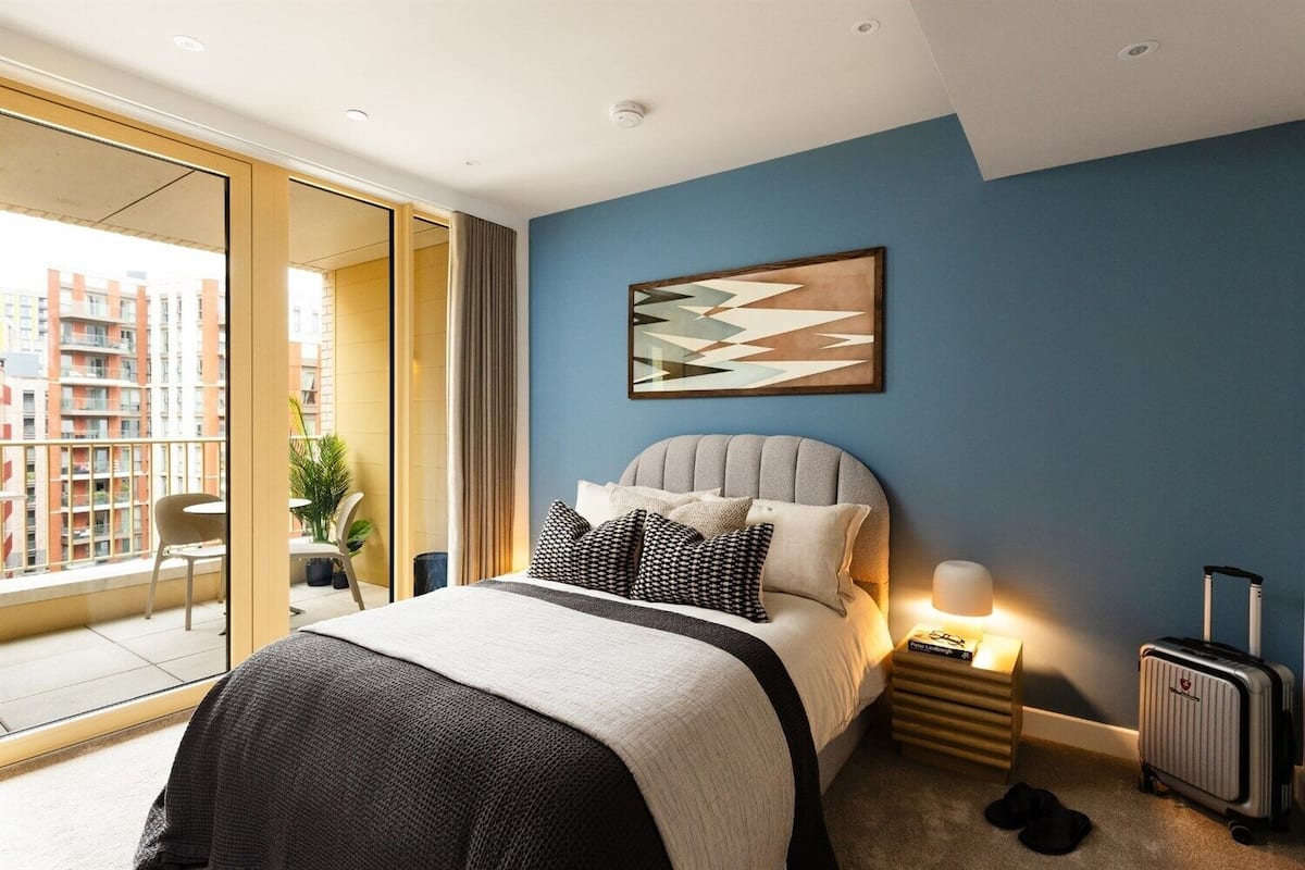 Un dormitorio moderno con una cama grande, una mesita de noche con lámpara, una maleta y vistas de la ciudad a través de una puerta corredera.