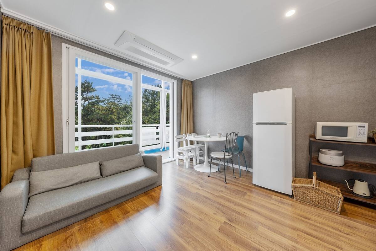 A modern living room with a grey sofa, a dining table with chairs, a white refrigerator, and a microwave.