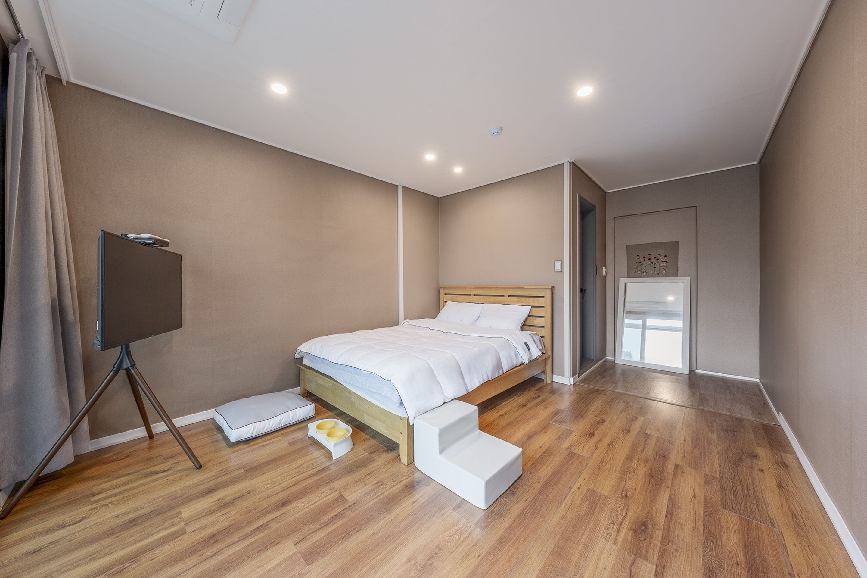 A modern bedroom with a wooden bed, a bedside table, a TV on a tripod stand, and a small step stool.