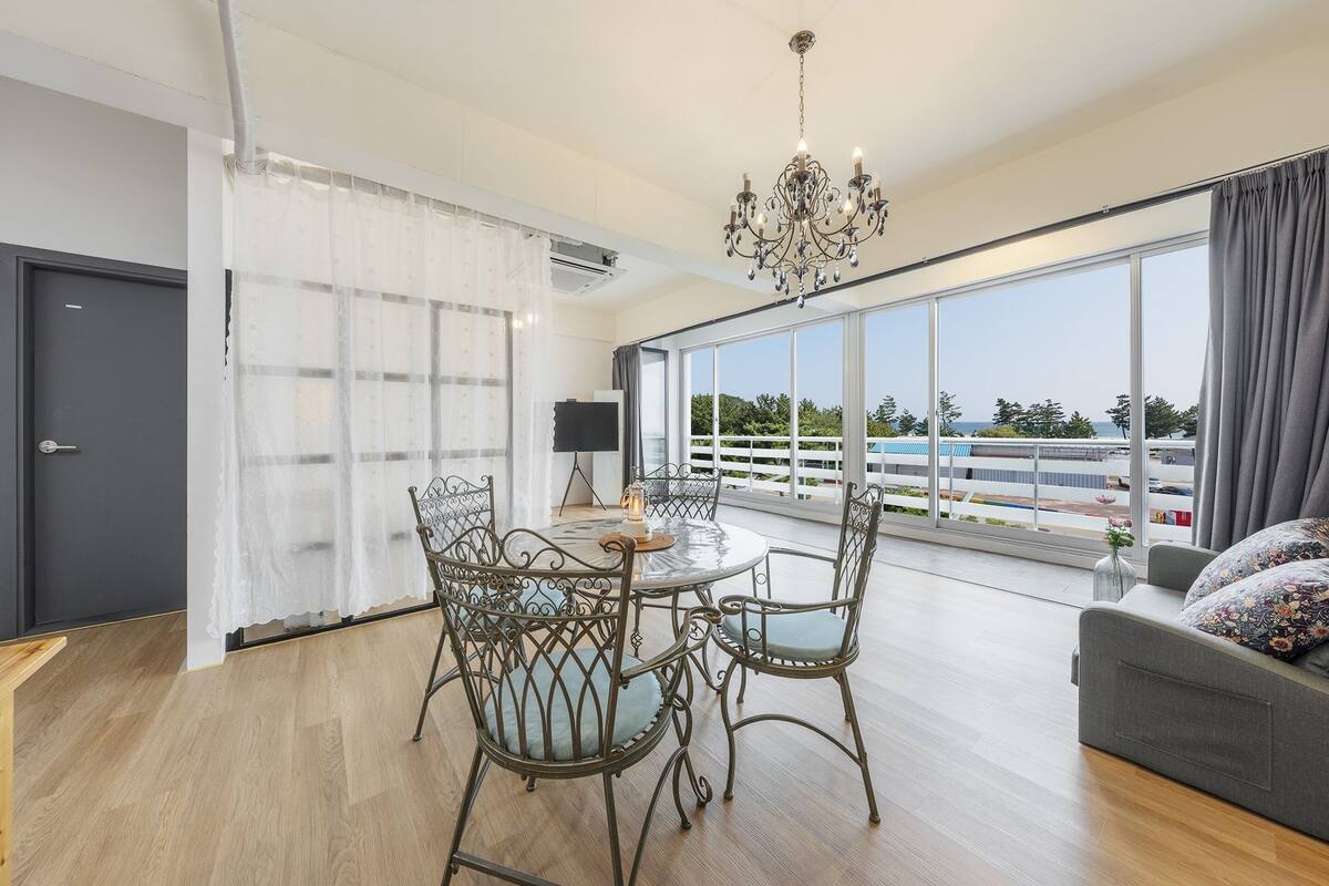 A modern living room with a round dining table, four chairs, a chandelier, a sofa, and large windows offering a view of the airport.