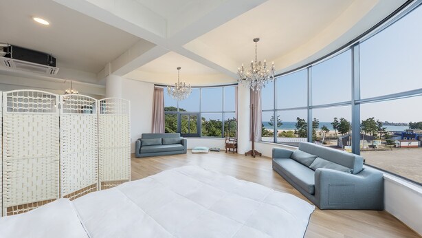 A spacious room with a large bed, two grey sofas, a white rug, and a view of the sea through large windows.