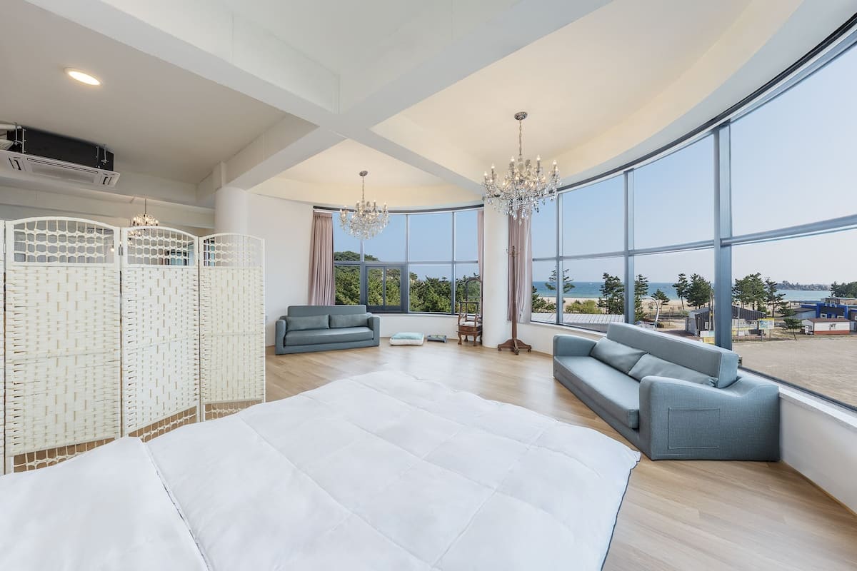 A spacious room with a large bed, two grey sofas, a white rug, and a view of the sea through large windows.