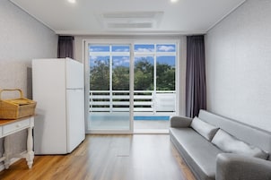 Un espacio habitable moderno y compacto con sofá, una mesita con una cesta, refrigerador y puerta corrediza de vidrio que da a un balcón exterior.