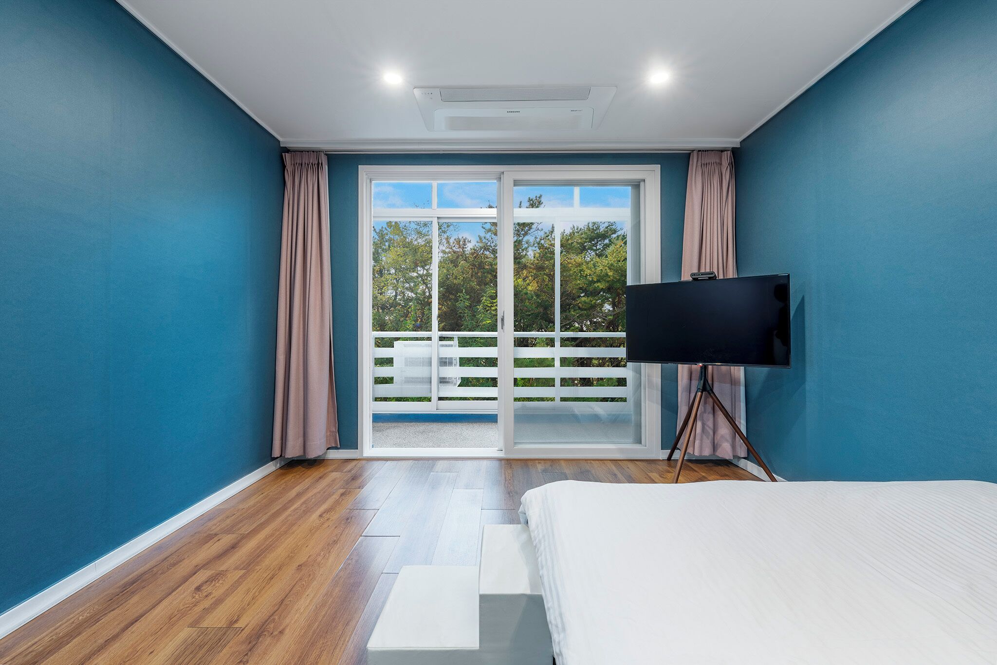 Un dormitorio con una cama, un televisor sobre un soporte de trípode y un ventanal corredero que da a un espacio exterior.