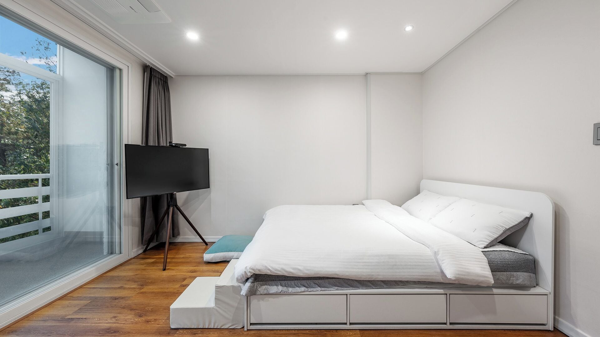 Un dormitorio moderno con cama, televisor sobre un mueble bajo y puerta corrediza de vidrio a un balcón.