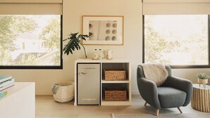 Uma sala de estar moderna com uma poltrona cinza, um pequeno armário branco e uma janela grande com vista para árvores e uma casa.