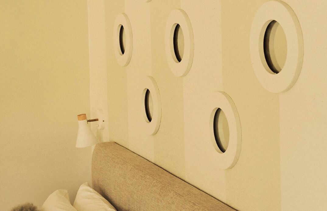 Una habitación con una cama, una pared con aberturas circulares y una pequeña mesa con una lámpara.