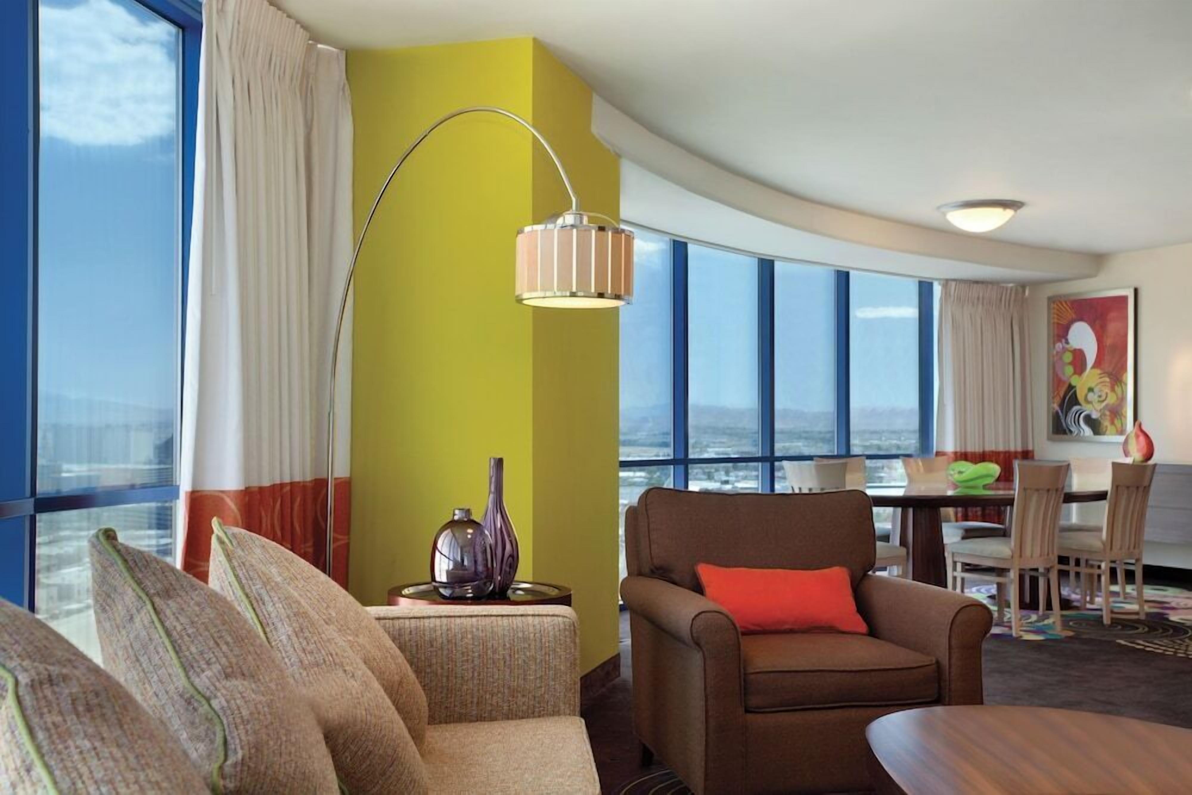 Ein modernes Hotelzimmer mit gewölbter Decke, einem großen Fenster mit Blick auf die Stadt, einer Essecke mit Tisch und Stühlen sowie einer Sitzecke mit Sofa und Sesseln.