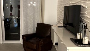 Une chambre moderne avec un fauteuil en cuir, un téléviseur à écran plat, un bureau avec une lampe et une fenêtre avec des rideaux.