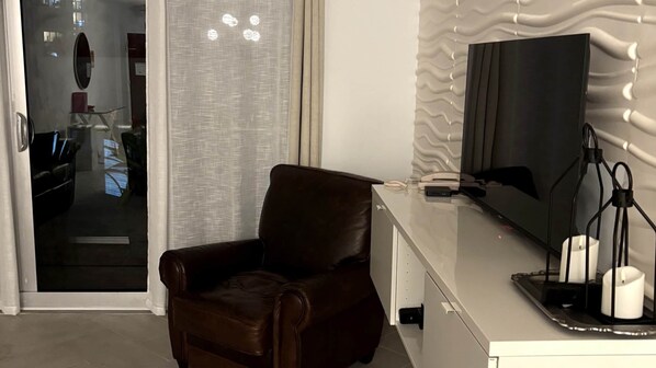 Ein modernes Zimmer mit Ledersessel, Flachbildfernseher, Schreibtisch und Lampe sowie einem Fenster mit Vorhängen.