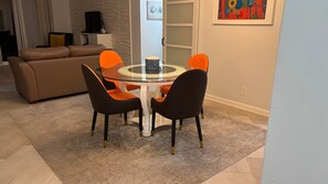 Un área de comedor con una mesa redonda de cristal y cuatro sillas, un sofá, un televisor y una obra de arte enmarcada en la pared.