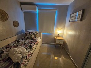 A small, well-lit room with a bed, a bedside table with a lamp, and an air conditioning unit. - Eduardo's Home (San Juan)